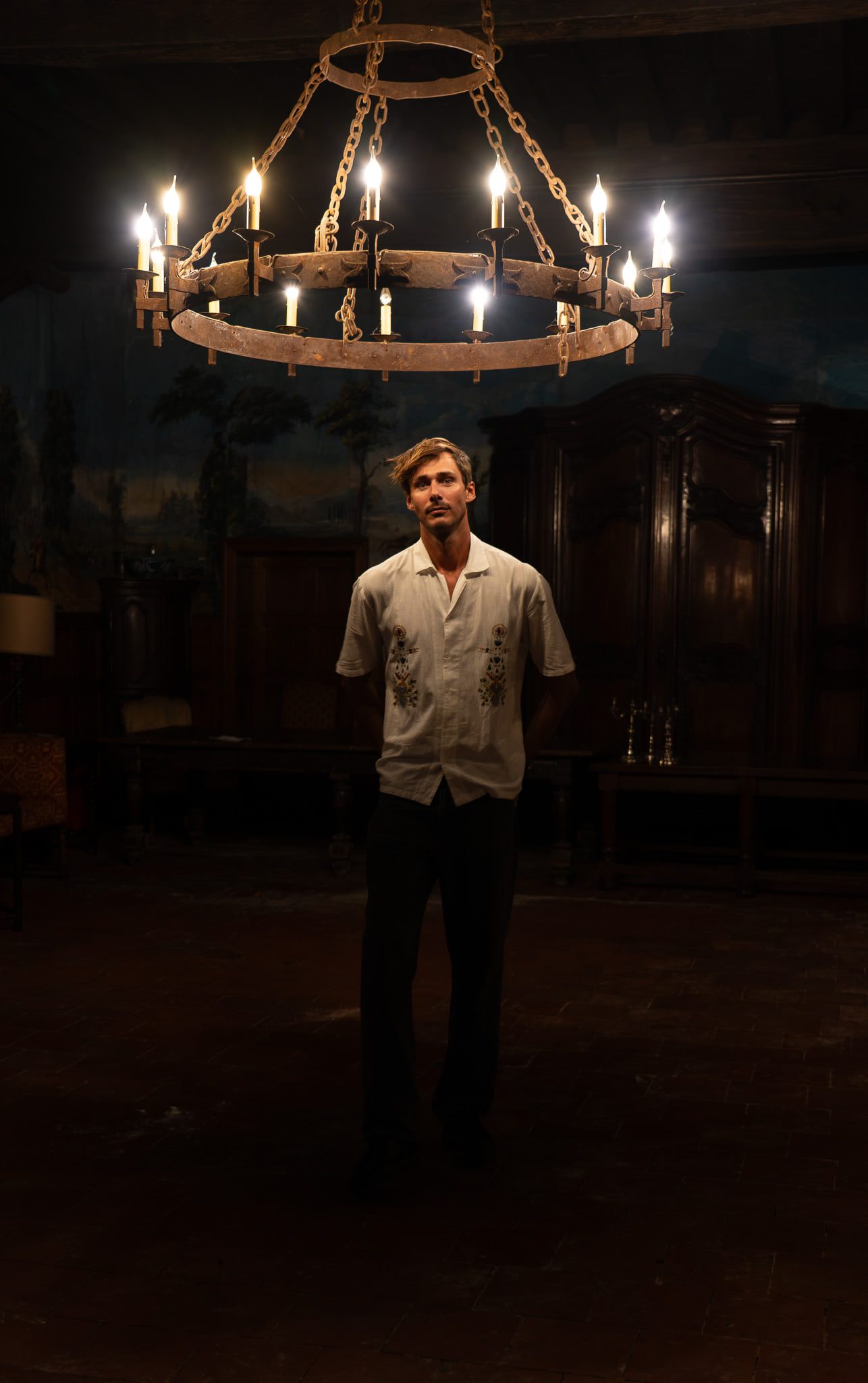 Victor Denver standing beneath a wrought-iron candle chandelier in a panelled château salon.