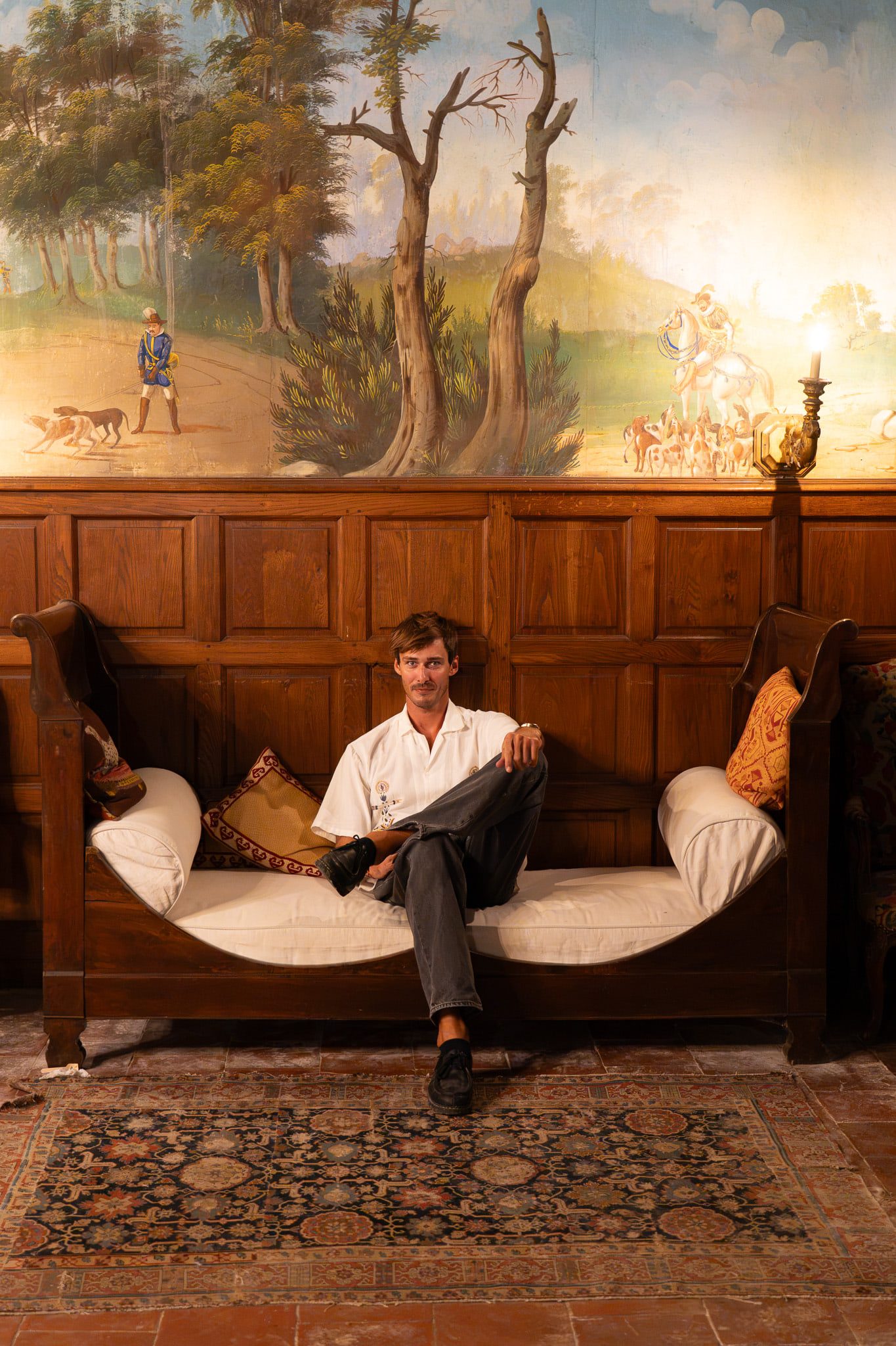 Victor Denver seated on a château daybed beneath a painted landscape mural, by candlelight.